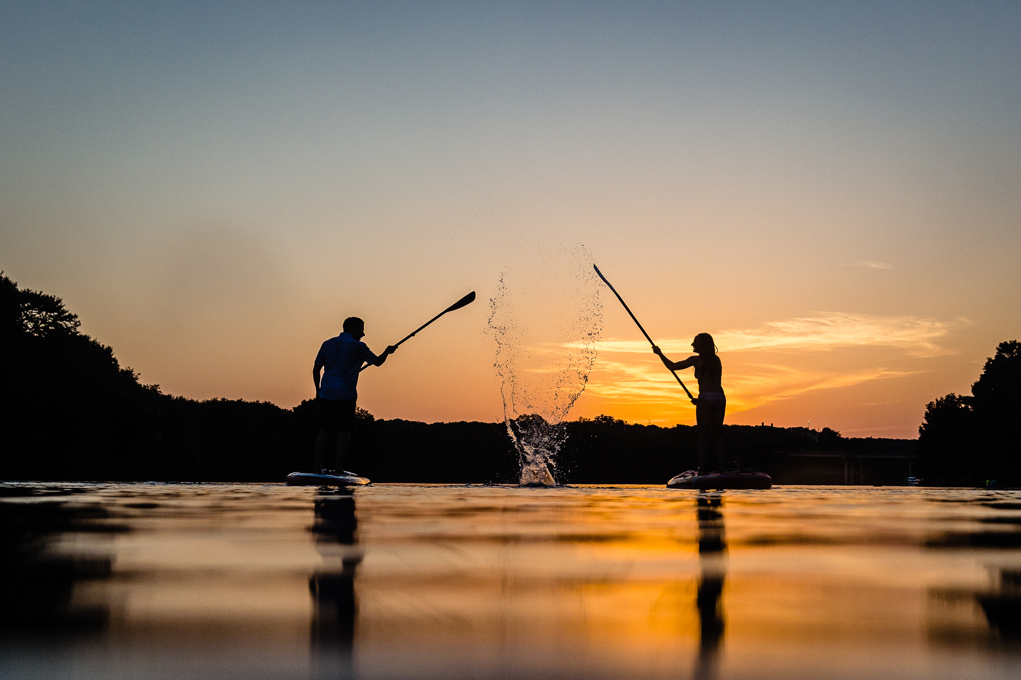 couple splashes each other with their paddles as they are silhouetted by the golden sunset